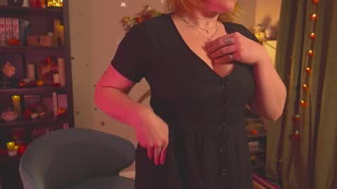 Media: Video of a plus-sized woman with fair skin, short red hair, wearing a black button-up dress, standing in a cozy room with shelves of books, warm lighting, and festive decorations.