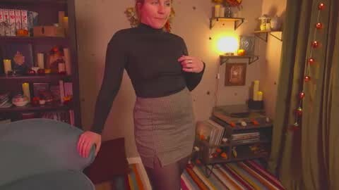 Media: Video of a smiling woman in a black turtleneck and high-waisted skirt, standing in a cozy, warmly lit room with shelves, books, candles, and a striped rug.