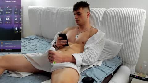 Media: Video of a young, shirtless, fair-skinned man with short dark hair, wearing white shorts and an unbuttoned white shirt, lying on a bed, playing with a vibrator on his penis, in a modern, white room.