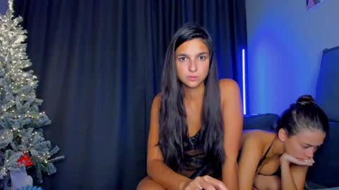 Media: A video featuring two young women in black lingerie sitting on a couch in a dimly lit room. One woman has long, straight black hair, and the other has brown hair in a ponytail. A Christmas tree with twinkling lights is visible in the background.
