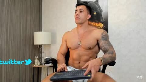 Media: Video of a shirtless, muscular, tattooed man with medium skin tone, short dark hair, wearing white shorts, typing on a keyboard in a modern room with a black panther poster and lamp.