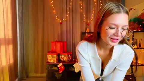 Media: Video of a smiling, fair-skinned, blonde woman with glasses, wearing a white cardigan, in a warmly lit, Christmas-decorated room with red and gold ornaments, gifts, and string lights.