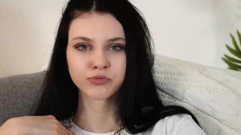 Media: A video of a young woman with fair skin, long black hair, and green eyes, sitting on a gray couch, wearing a white top and a gold necklace. The background is blurred, showing a white wall and a potted plant.