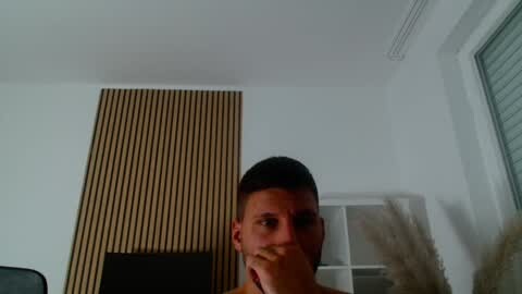 Media: Video of a young man with short dark hair, partially obscured by a finger, standing in a minimalist room with a wooden-paneled wall, a window with blinds, and a beige curtain.