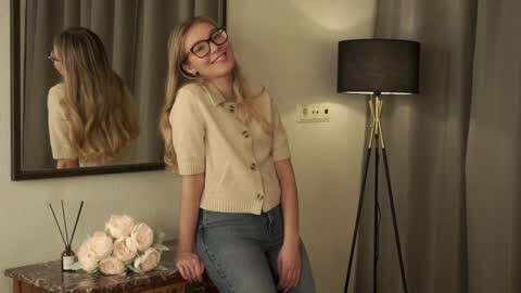 Media: Video of a young woman with long blonde hair, wearing glasses, a beige blouse, and jeans, leaning against a table with flowers, in a dimly lit room with a black floor lamp.