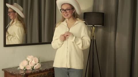 Media: Video of a smiling Caucasian woman in a white cowboy hat, glasses, and cream jacket, standing in a room with a mirror, flowers, and a floor lamp.