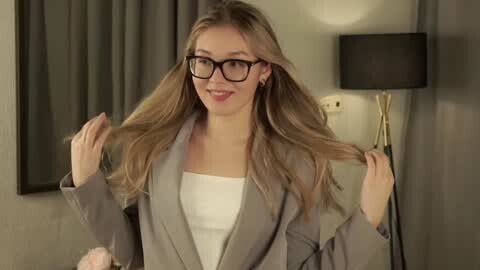 Media: Video of a young Caucasian woman with fair skin, long blonde hair, and glasses, wearing a gray blazer over a white top, smiling, in a modern hotel room with beige walls and a black lamp.