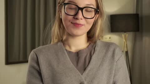 Media: Video of a fair-skinned woman with blonde hair, wearing black-rimmed glasses, a gray sweater, and a gray shirt, smiling softly, in a modern room with a black lamp, beige curtains, and a neutral background.