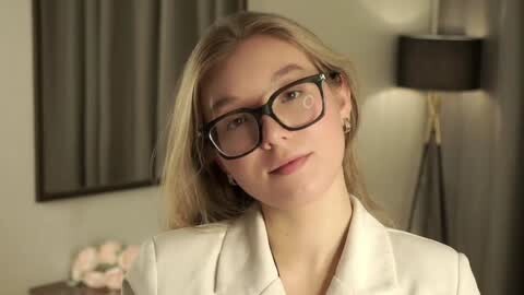 Media: Video of a fair-skinned, blonde woman with glasses, wearing a white blazer, smiling slightly. Background features a modern lamp, gray curtains, and a framed picture.