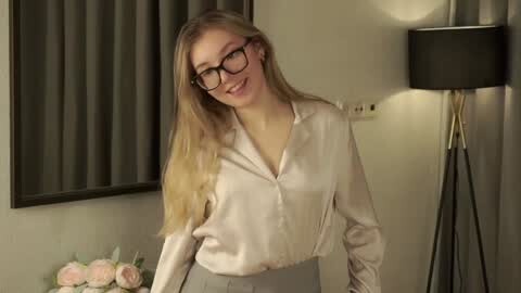 Media: Video of a smiling, fair-skinned woman with long blonde hair, wearing glasses, a white blouse, and grey skirt, standing in a modern room with grey curtains, a lamp, and a vase of pink roses.
