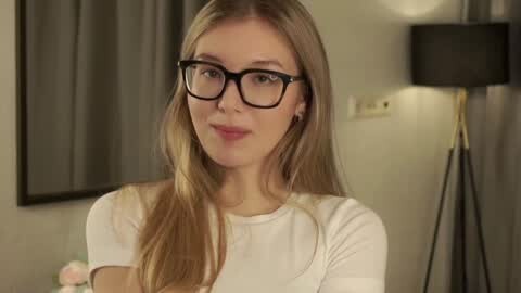 Media: Video of a young, fair-skinned woman with long, straight blonde hair, wearing black-rimmed glasses and a white top, standing indoors with beige walls, a lamp, and a gray curtain in the background.