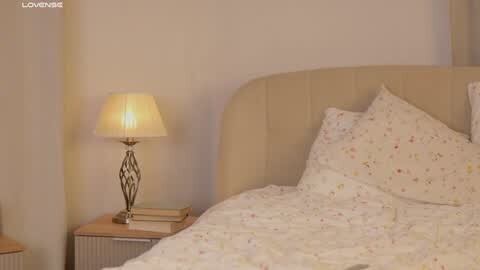 Media: A video of a cozy bedroom with a beige upholstered headboard, a white bedspread adorned with small floral patterns, and a wooden nightstand with a lamp featuring a pleated lampshade.