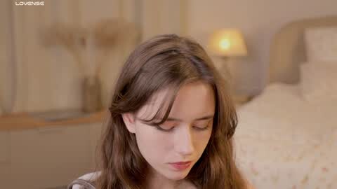 Media: A video of a young woman with long brown hair and fair skin, sitting on a bed in a softly lit, cozy bedroom with a beige headboard and a warm lamp.