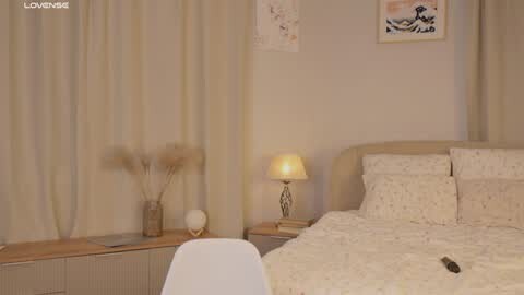 Media: Video of a serene, beige-toned bedroom with a neatly made bed, soft lighting, and minimalist decor including a lamp, vase, and chair.