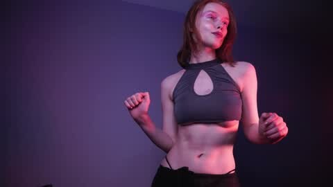 Media: Video of a fit, fair-skinned woman with long red hair, wearing a black sports bra and black leggings, striking a boxing pose against a dark, purple-lit background.