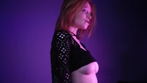 Media: Video of a fair-skinned, red-haired woman in a black, fishnet top, standing against a purple backdrop, showcasing her slender physique.