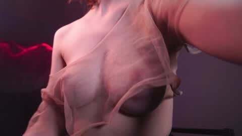 Media: Video of a woman with fair skin, wearing a sheer, see-through pink top, revealing her medium-sized breasts and nipples. The background is dimly lit with a red hue, creating a sensual atmosphere.