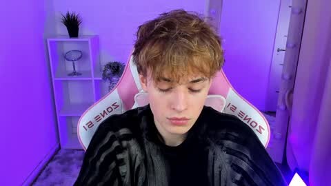Media: Video of a young, fair-skinned boy with curly brown hair, wearing a black shirt, sitting on a pink gaming chair with white text, in a dimly lit room with purple lighting.
