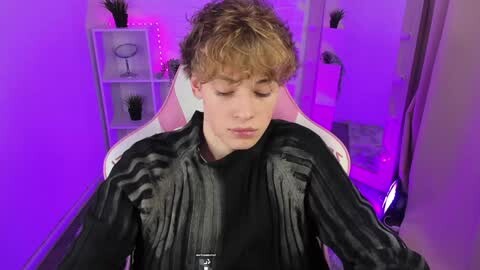 Media: Video of a young man with curly blonde hair, wearing a black leather jacket, seated in a pink chair with purple lighting in a minimalist room.