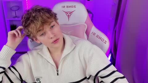 Media: Video of a young, fair-skinned, curly-haired boy with a slender build, wearing a white ribbed sweater, seated in a white gaming chair with \"Xbox\" branding. Background features purple lighting and a white shelf with a mirror.