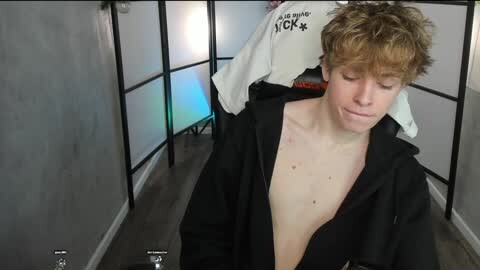 Media: A video of a young, shirtless, fair-skinned boy with messy blonde hair, wearing a black hoodie, standing in a dimly lit room with grey walls and a white \"NICK STAR\" sweatshirt hanging nearby.