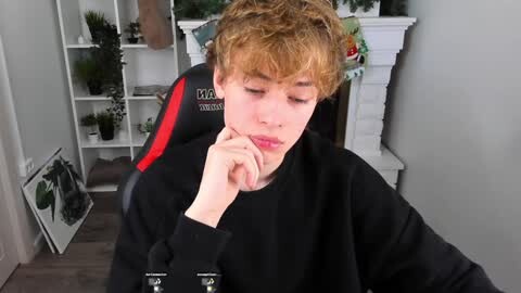 Media: Video of a young, fair-skinned boy with curly, light brown hair, wearing a black shirt, sitting in a black and red gaming chair, deep in thought, in a modern living room with white shelves and green plants.