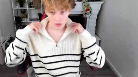 Media: Video of a young boy with light skin and messy blonde hair, wearing a white and black striped sweater, adjusting the zipper. Background shows a white fireplace with a green wreath and white shelves with plants and decor.