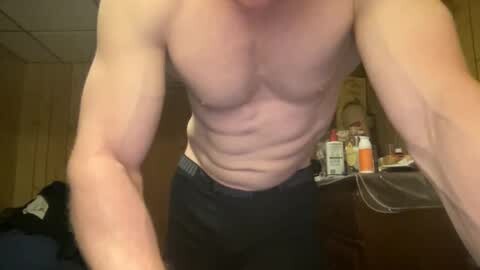 Media: Video of a shirtless, muscular man with fair skin and black pants, standing in a dimly lit room with wooden paneling. Background includes a cluttered desk with various items.