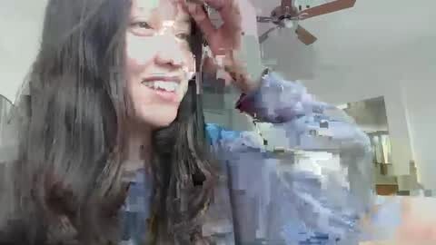 Media: A video of a young woman with long, dark hair and a slight smile, standing in a room with a ceiling fan and a partially visible person in the background.