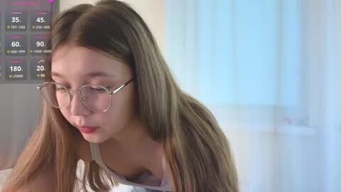 Media: Video of a young woman with long, straight brown hair, wearing glasses, sitting at a desk, looking down. Background includes a digital screen displaying weather data.