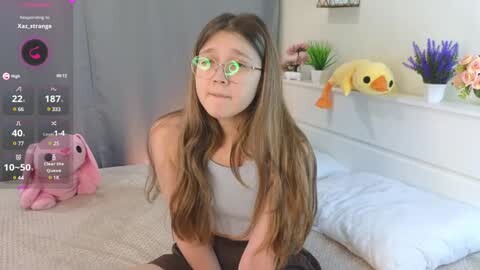 Media: A video of a young woman with long brown hair and green glasses, sitting on a bed with plush toys. The room has a cozy, pastel-colored decor with flowers and a digital clock.