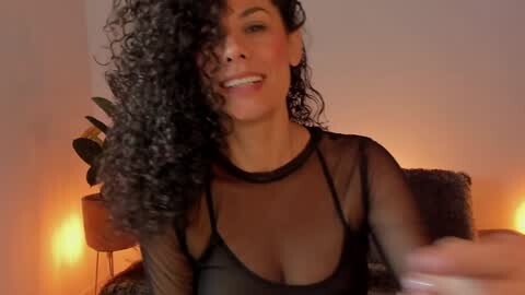 Media: Video of a smiling, curly-haired woman with medium brown skin, wearing a black sheer top, holding a black curly wig. Warm, dimly lit room with potted plants in the background.