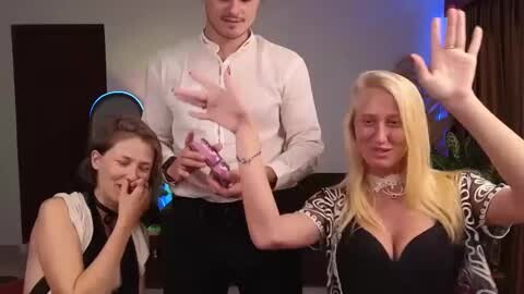 Media: Video of three people: a man with a white shirt, a woman with long blonde hair in a black dress, and a young woman with a headset, all engaged in a playful, intimate moment.