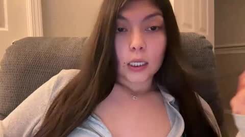 Media: Video of a young Asian woman with long brown hair, wearing a light blue top and a gray cardigan, seated on a gray couch in a living room.