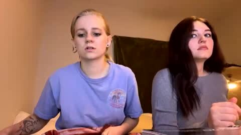 Media: Video of two young women, one blonde in a blue shirt, the other brunette in a gray shirt, seated at a table in a dimly lit room, discussing something intensely.