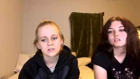 Media: Video of two young women with light skin, one with blonde hair in a ponytail and the other with dark hair, both wearing casual black hoodies, sitting on a bed with green drapes in the background.