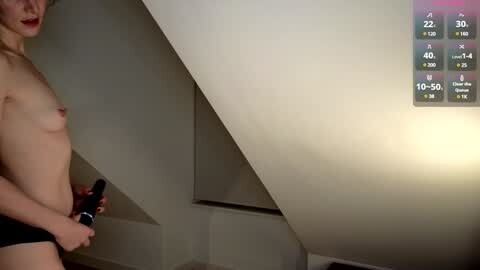 Media: Video of a slender, fair-skinned woman with small breasts, topless, wearing black shorts, holding a black object, standing in a dimly lit attic with a digital display showing exercise stats on the wall.