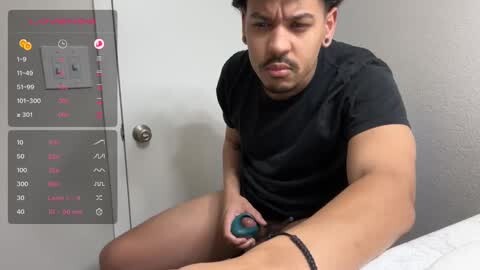 Media: Video of a young Asian man with short dark hair, wearing a black shirt and shorts, sitting on a bed, using a blue vibrator on his left leg, while a digital display shows temperature and intensity settings.