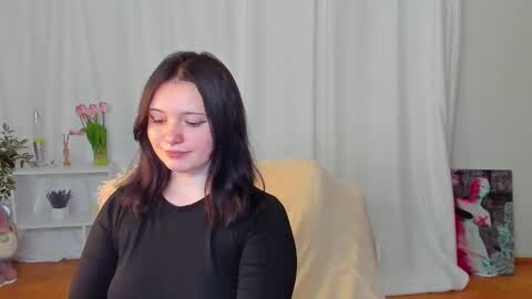 Media: Video of a young woman with long, dark hair, wearing a black top, seated in a well-lit room with white curtains, a wooden table, and a potted plant.