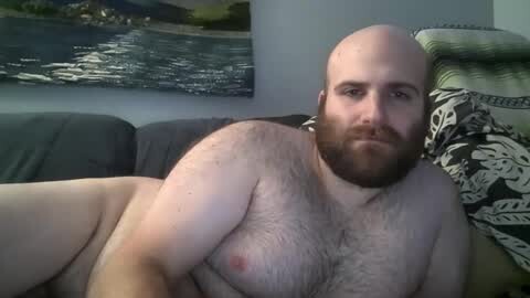 Media: Video of a bald, bearded, overweight, light-skinned man, lying nude on a couch, with a framed landscape painting and a patterned pillow in the background.