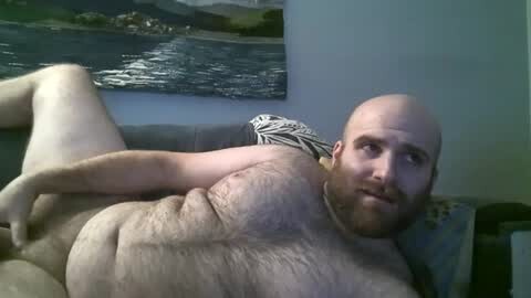 Media: Video of a bald, bearded, naked man with a medium build, lying on a couch. He has fair skin and is relaxed, with a neutral expression. The background features a framed painting of a coastal scene and a patterned pillow.