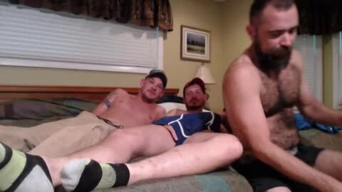 Media: Video of three shirtless, bearded men in underwear on a bed, one man holding a cellphone, with a framed landscape painting on the wall.