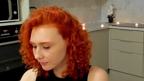 Media: Video of a young woman with voluminous, curly red hair, wearing a black top, standing in a modern kitchen with beige cabinets, white marble countertops, and candles.