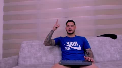 Media: Video of a tattooed man with short dark hair, wearing a blue Nike T-shirt, sitting on a light grey couch, pointing with his index finger, holding a remote control.