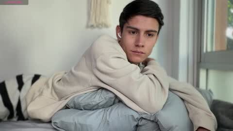 Media: Video of a young man with short dark hair, wearing a cream sweater and light blue pants, lying on a bed with a beige blanket, looking thoughtful in a softly lit room.