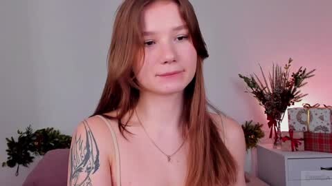 Media: Video of a young, fair-skinned woman with long brown hair, wearing a beige tank top, standing indoors. She has a small tattoo on her left shoulder. Background includes green potted plants, a lit lamp, and a red gift box.