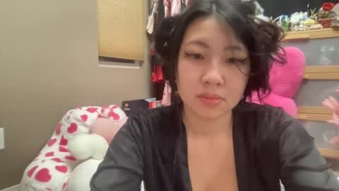 Media: Video of a young Asian woman with curly black hair, wearing a black robe, sitting in a cluttered room with pink furniture and stuffed animals.