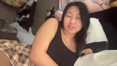 Media: Video of an East Asian woman with long black hair, medium build, wearing a black tank top, lying on a bed in a cluttered bedroom.