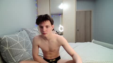Media: Video of a young, slim, shirtless man with short dark hair, wearing black underwear, sitting on a bed with a gray geometric-patterned pillow, in a modern bedroom with a wooden wardrobe and a ring light.