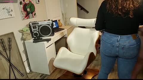 Media: Video of a woman with curly hair, wearing a black top and blue jeans, adjusting a white Eames chair in a modern, cozy room with wooden floors, a white desk, and colorful artwork on the walls.